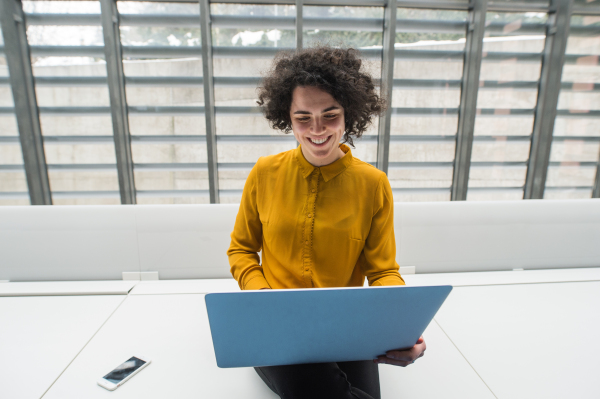 A portrait of a young student or businesswoman sitting on desk in room in a library or office, using laptop.