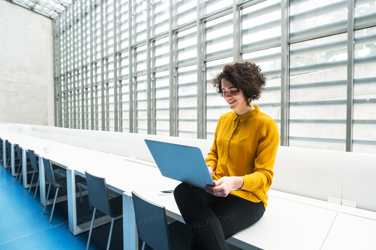 A portrait of a young student or businesswoman sitting on desk in room in a library or office, using laptop.