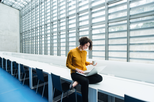 A portrait of a young student or businesswoman sitting on desk in room in a library or office, using laptop.