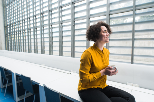 A portrait of a young student or businesswoman sitting on desk in room in a library or office, holding coffee.