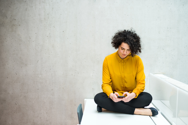 Portrait of a young student or businesswoman with smartphone sitting on desk in room in a library or office. Copy space.