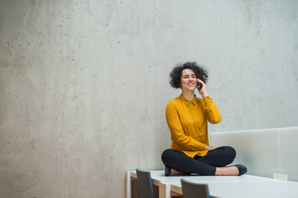 Young student or businesswoman sitting cross-legged on desk in room in a library or office, using smartphone. Copy space.