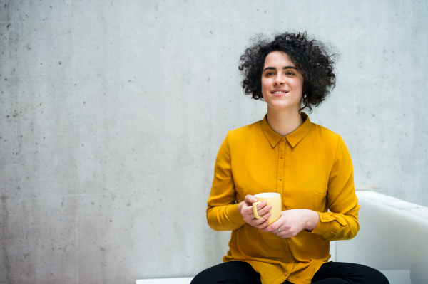 A portrait of a young student or businesswoman sitting on desk in room in a library or office, holding coffee.