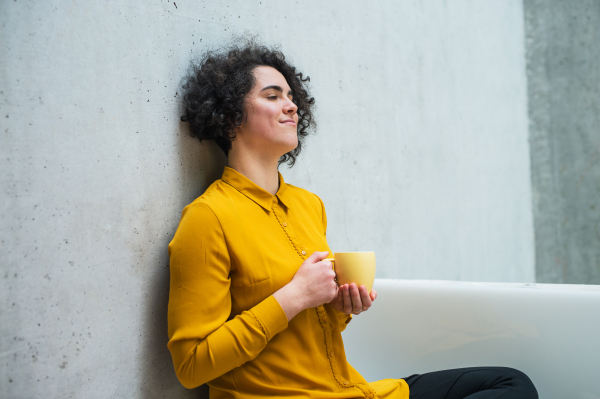 A portrait of a young student or businesswoman sitting on desk in room in a library or office, holding coffee.