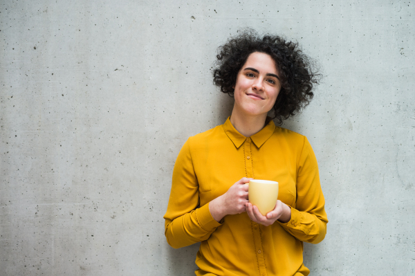 A portrait of young cheerful businesswoman standing in office, holding a cup of coffee.