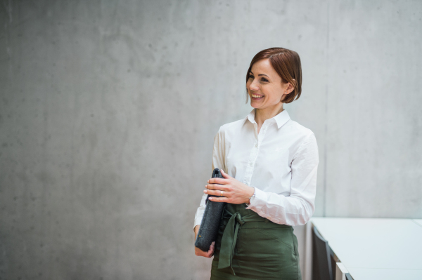 A portrait of young cheerful businesswoman standing in office, holding laptop in a bag.