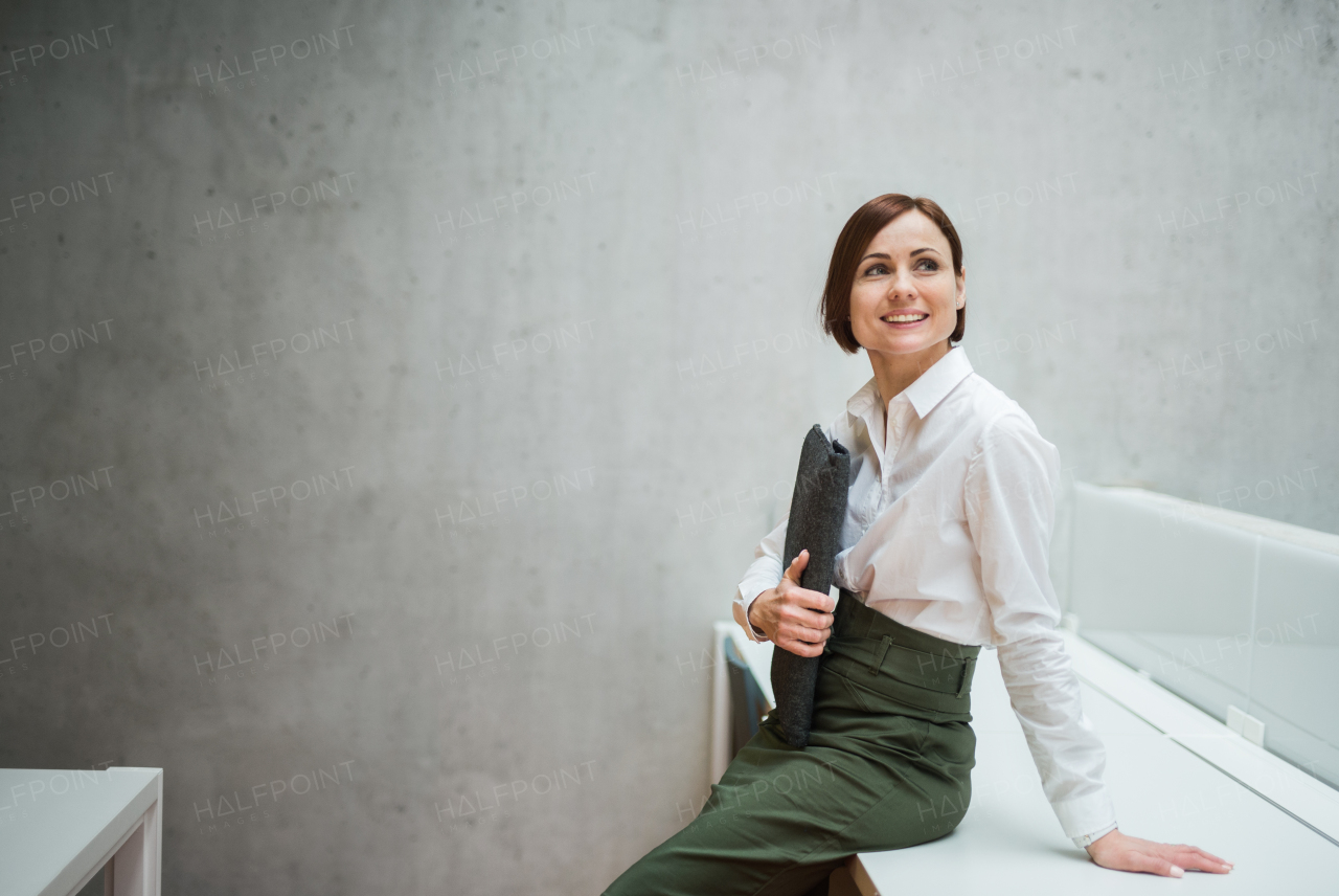 A portrait of young cheerful businesswoman sitting on desk in office, holding laptop in a bag.