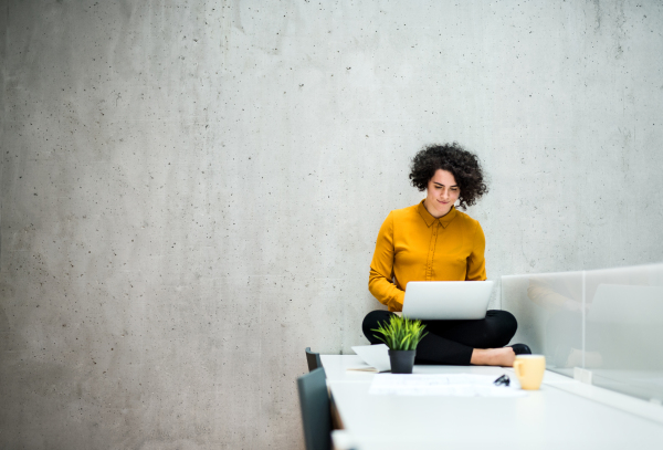 A portrait of a young student or businesswoman sitting on desk in room in a library or office, using laptop.