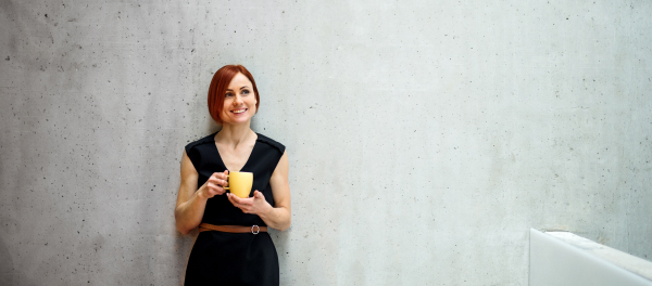 A portrait of young cheerful businesswoman standing in office, holding a cup of coffee.