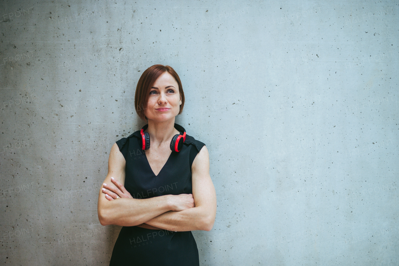 A portrait of young business woman with headphones standing against concrete wall in office, arms crossed.