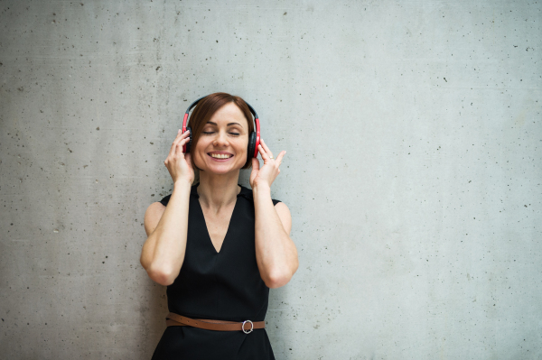 A portrait of young business woman with headphones standing against concrete wall in office and listening to music, eyes closed.