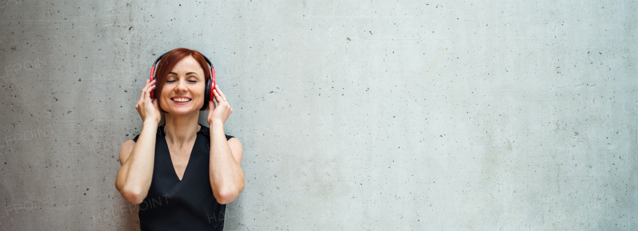 A portrait of young business woman with headphones standing against concrete wall in office and listening to music.