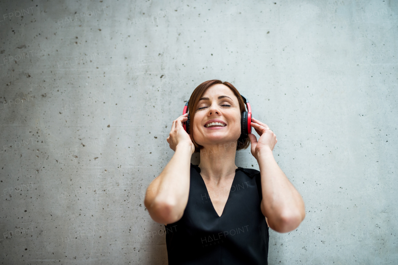 A portrait of young business woman with headphones standing against concrete wall in office and listening to music, eyes closed.