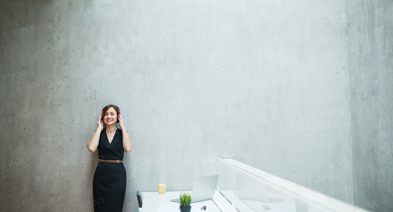 A portrait of young business woman with headphones standing against concrete wall in office and listening to music.