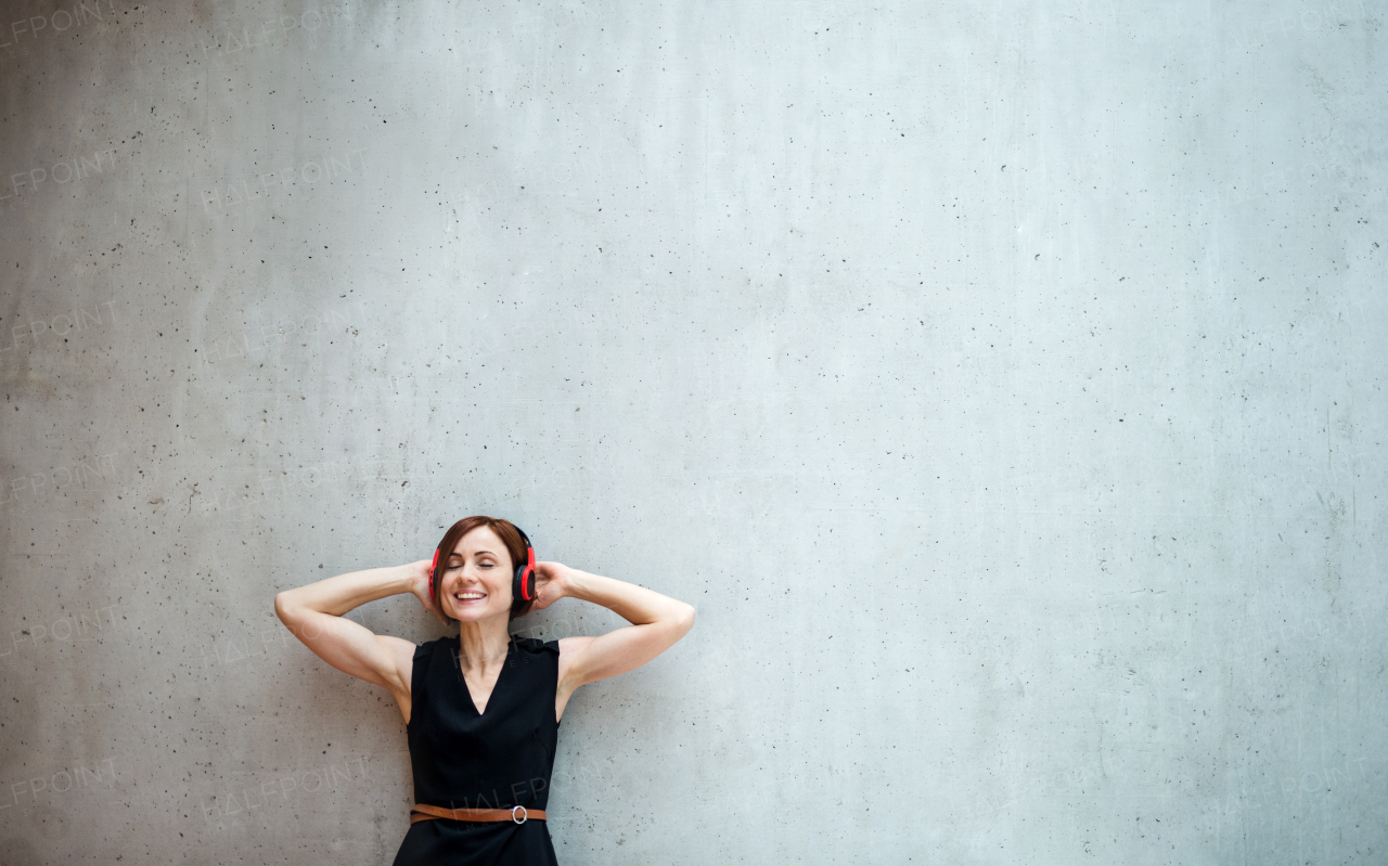 A portrait of young business woman with headphones standing against concrete wall in office and listening to music.