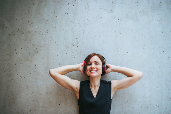 A portrait of young business woman with headphones standing against concrete wall in office and listening to music, eyes closed.