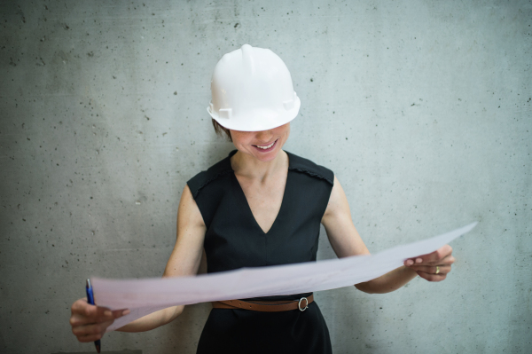 A young businesswoman or architect with white helmet standing in office, looking at blueprints.