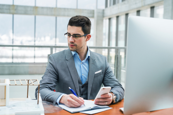 Young businessman or architect sitting at the desk with computer and smartphone in office, looking at house model and working.