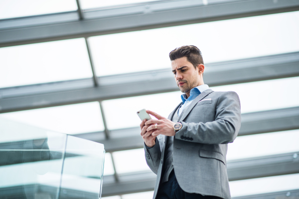 A low-angle view of young serious businessman with smartphone in corridor outside office