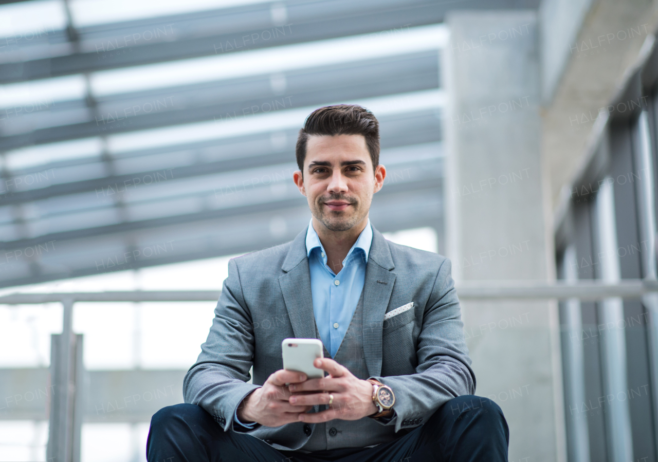 A portrait of happy young businessman with smartphone sitting in corridor outside office.