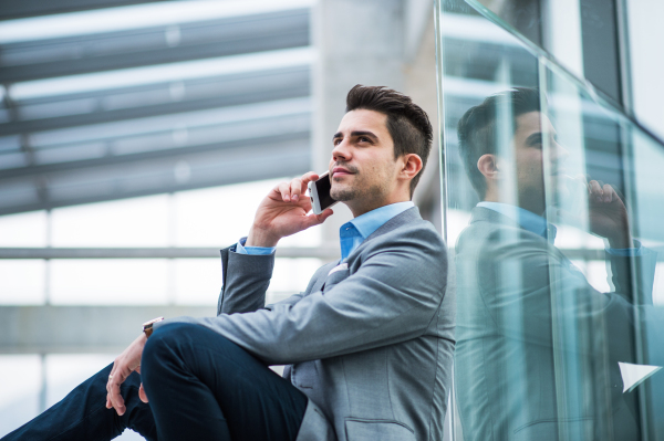 A portrait of happy young businessman with smartphone sitting in corridor outside office, making a phone call.