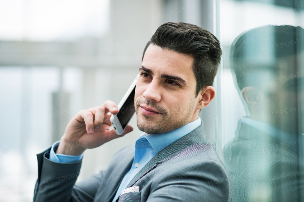 A portrait of happy young businessman with smartphone sitting in corridor outside office, making a phone call.