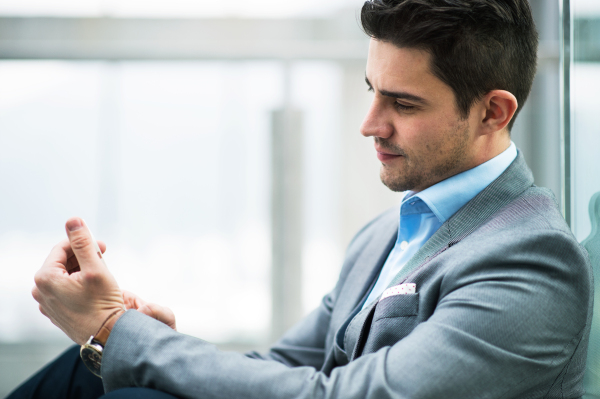 A portrait of happy young businessman sitting in corridor outside office, using smartphone.