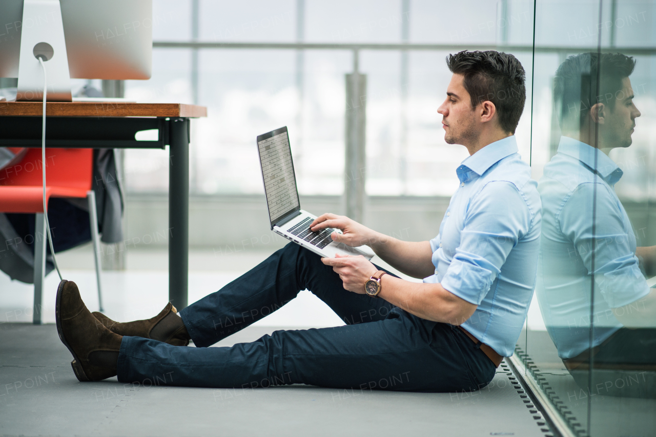 A portrait of young businessman with laptop sitting on the floor in office, working.