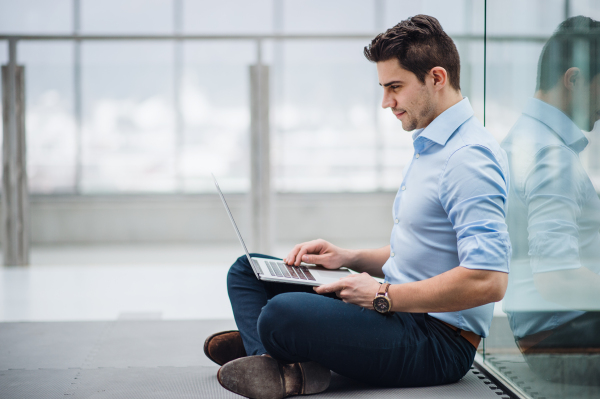 A portrait of young businessman with laptop sitting on the floor in corridor outside office.