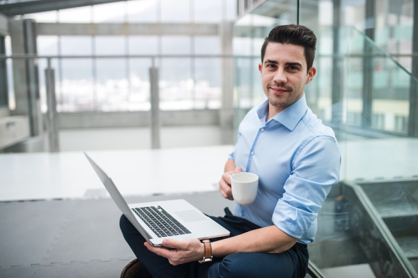 A portrait of young businessman with laptop and coffee sitting on the floor in corridor outside office.