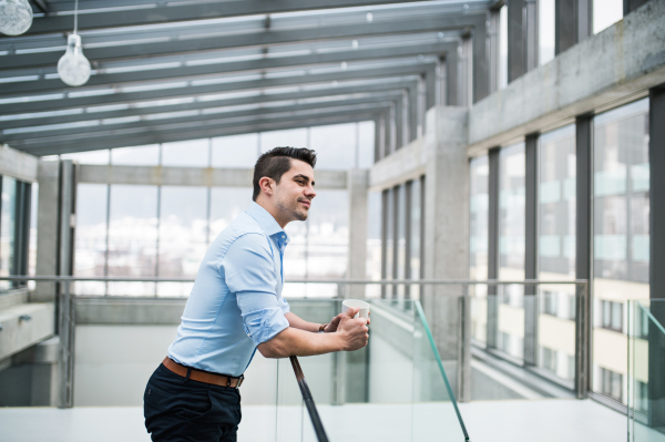 A side view of young businessman with cup of coffee standing in corridor outside office, resting.