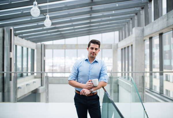 A young businessman with cup of coffee standing in corridor outside office, looking at camera.