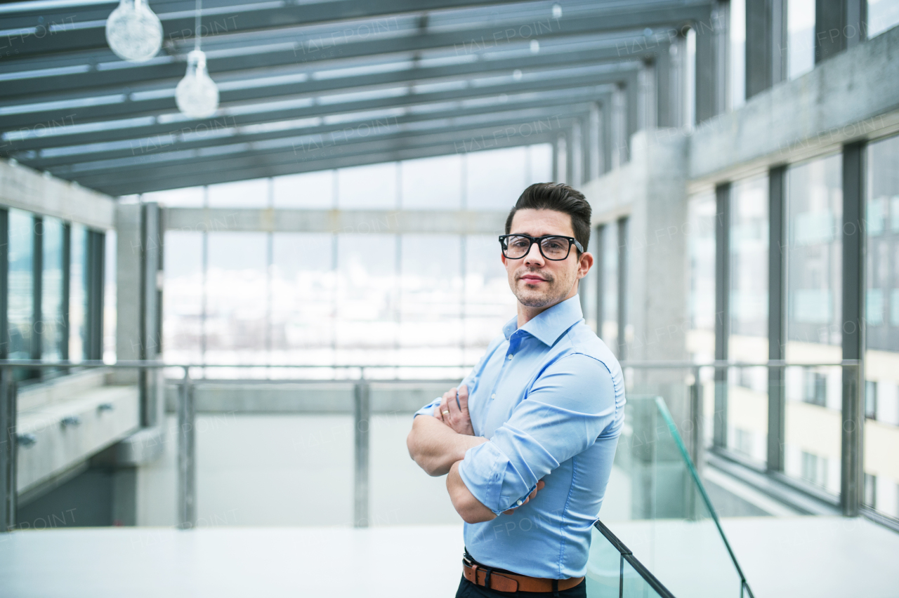 A portrait of young businessman standing indoors in an office, arms crossed. Copy space.