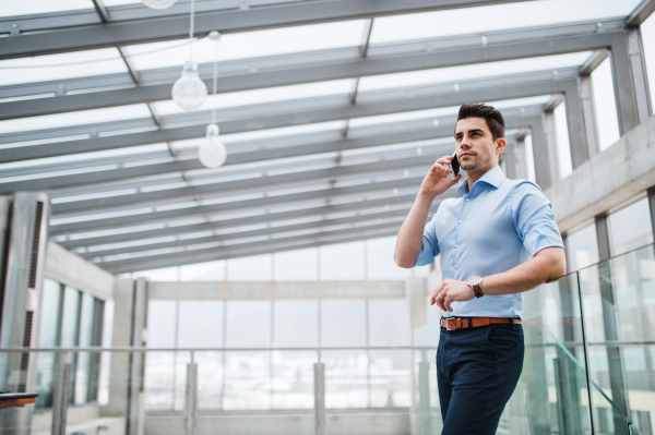 A low-angle view of young serious businessman with smartphone in corridor outside office, making a phone call.