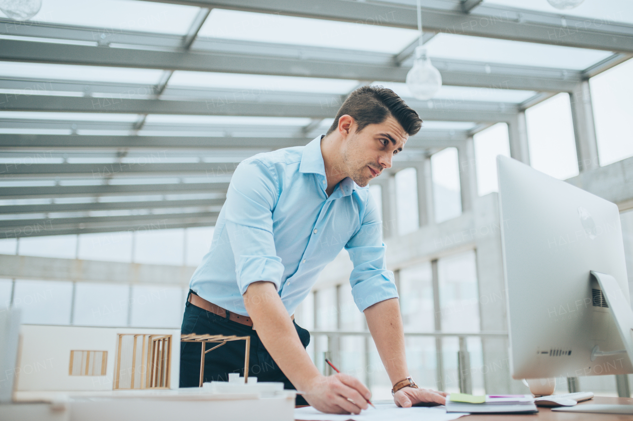 Young businessman or architect with model of a house standing at the desk in office, working.