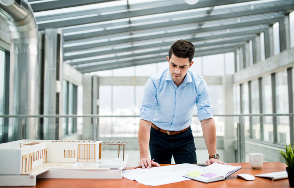 Young businessman or architect with model of a house standing at the desk in office, working.