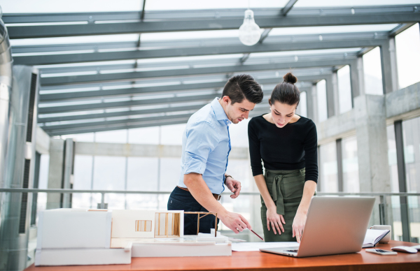 Two young male and female architects with laptop, blueprints and model of a house standing in office, talking.