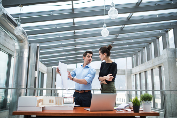 Two young male and female architects with laptop, blueprints and model of a house standing in office, talking.