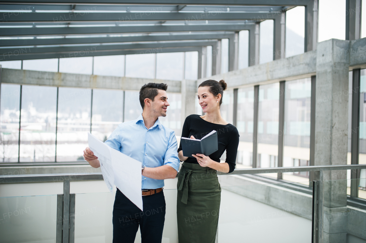 Two young male and female architects with blueprints standing in office, talking.