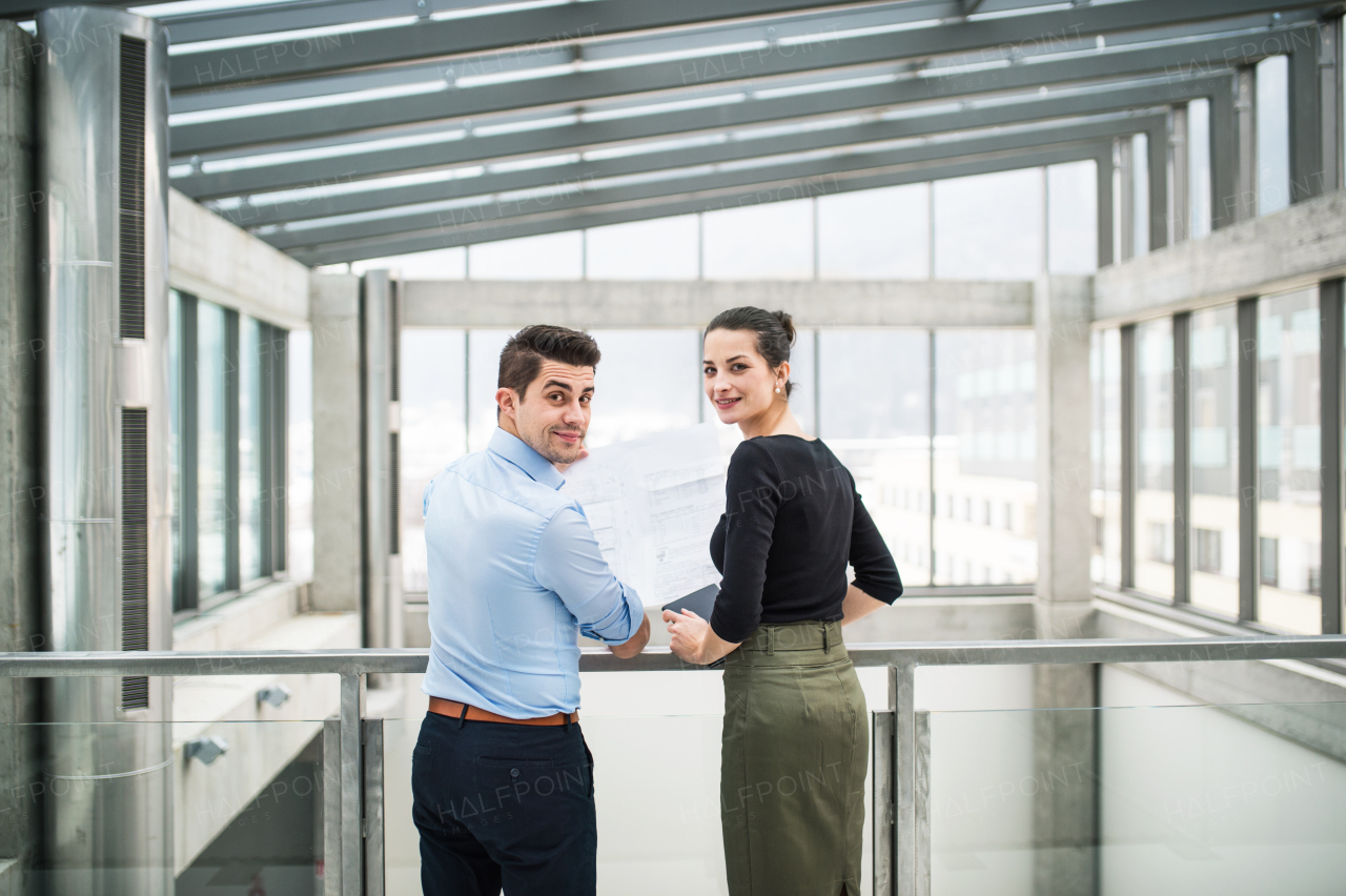 A rear view of two young male and female architects with blueprints standing in office, looking back.