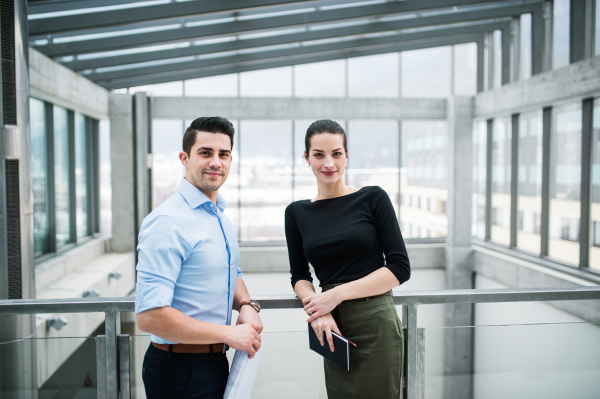 Two young male and female architects with blueprints standing in office, looking at camera.