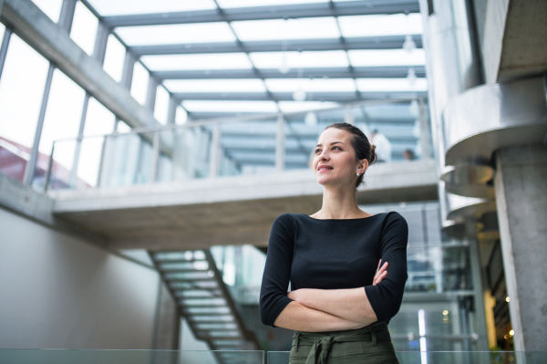 A waist-up portrait of young businesswoman standing in corridor outside office, arms crossed.