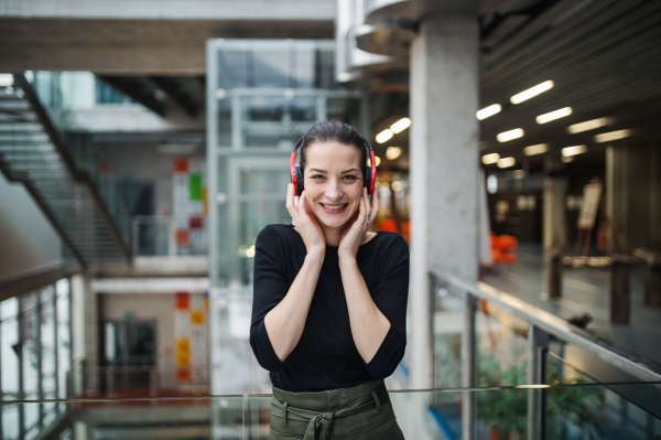 A portrait of happy young businesswoman with headphones standing in corridor outside office, listening to music.