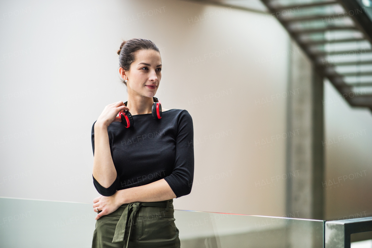 A portrait of happy young businesswoman with headphones standing in corridor outside office.