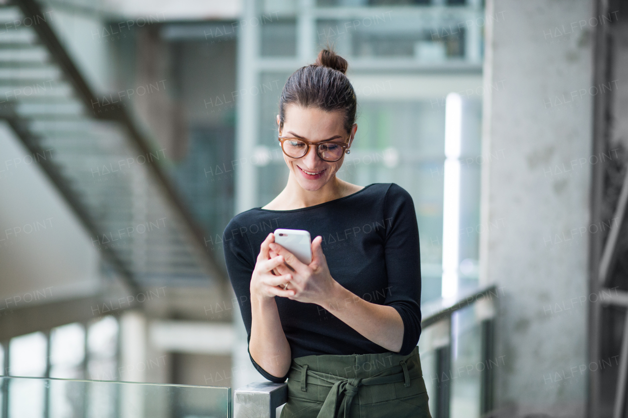 A portrait of happy young businesswoman with glasses and smartphone standing in corridor outside office.