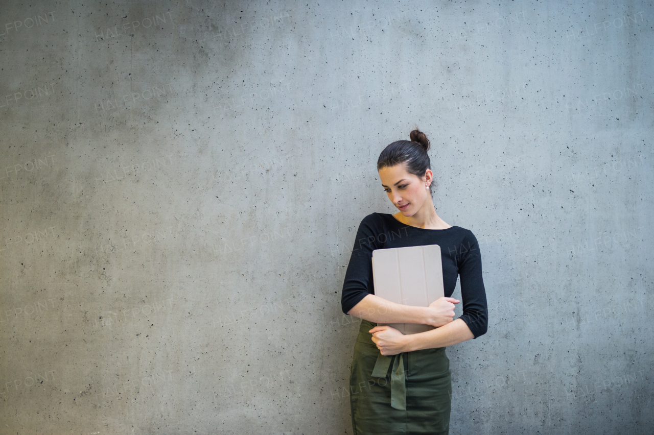 A portrait of happy young business woman with digital tablet standing against concrete wall in office. Copy space.