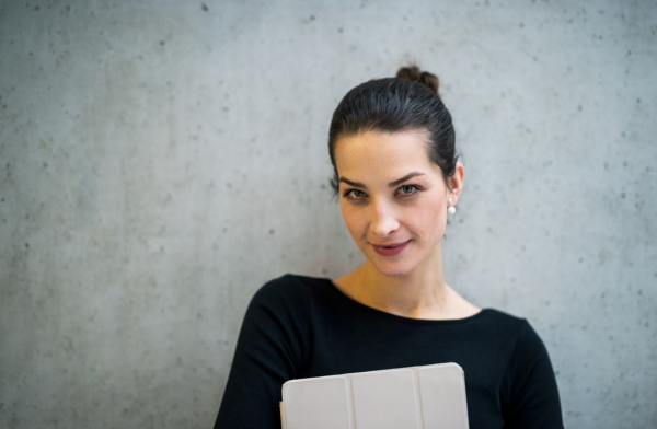 A portrait of happy young business woman with digital tablet standing against concrete wall in office.