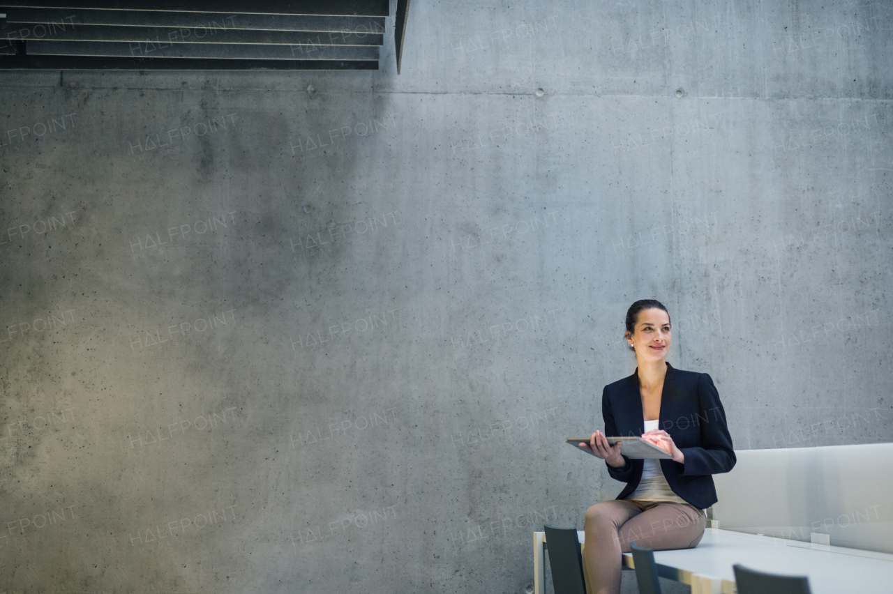 Young business woman with tablet sitting on desk against concrete wall in office. Copy space.