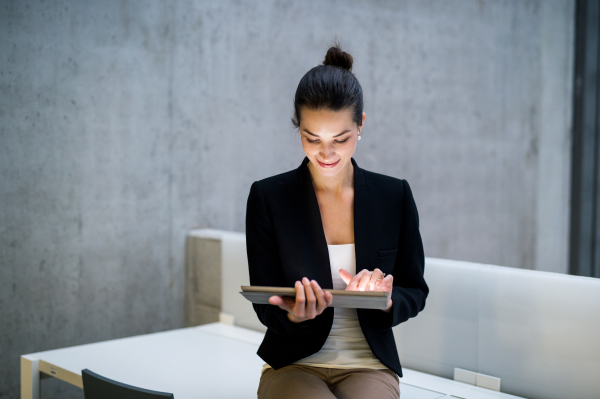 A portrait of a young student or businesswoman sitting on desk in room in a library or office, using tablet.