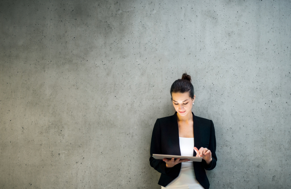 A portrait of happy young business woman with digital tablet standing against concrete wall in office.